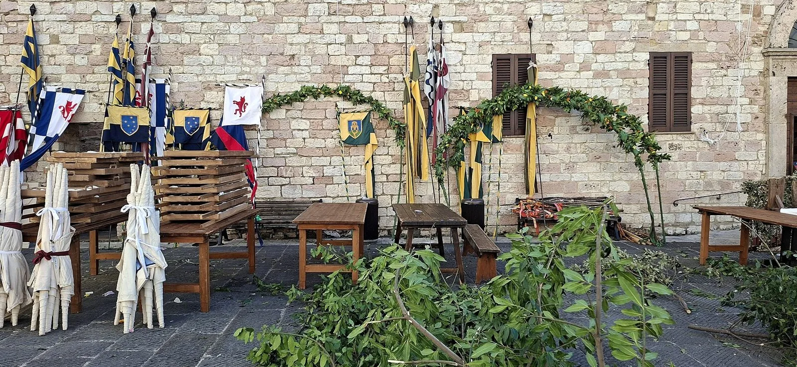 Stendardi e vessilli delle Parti appoggiati a un muro in pietra di Assisi, tra tavoli di legno e rami verdi